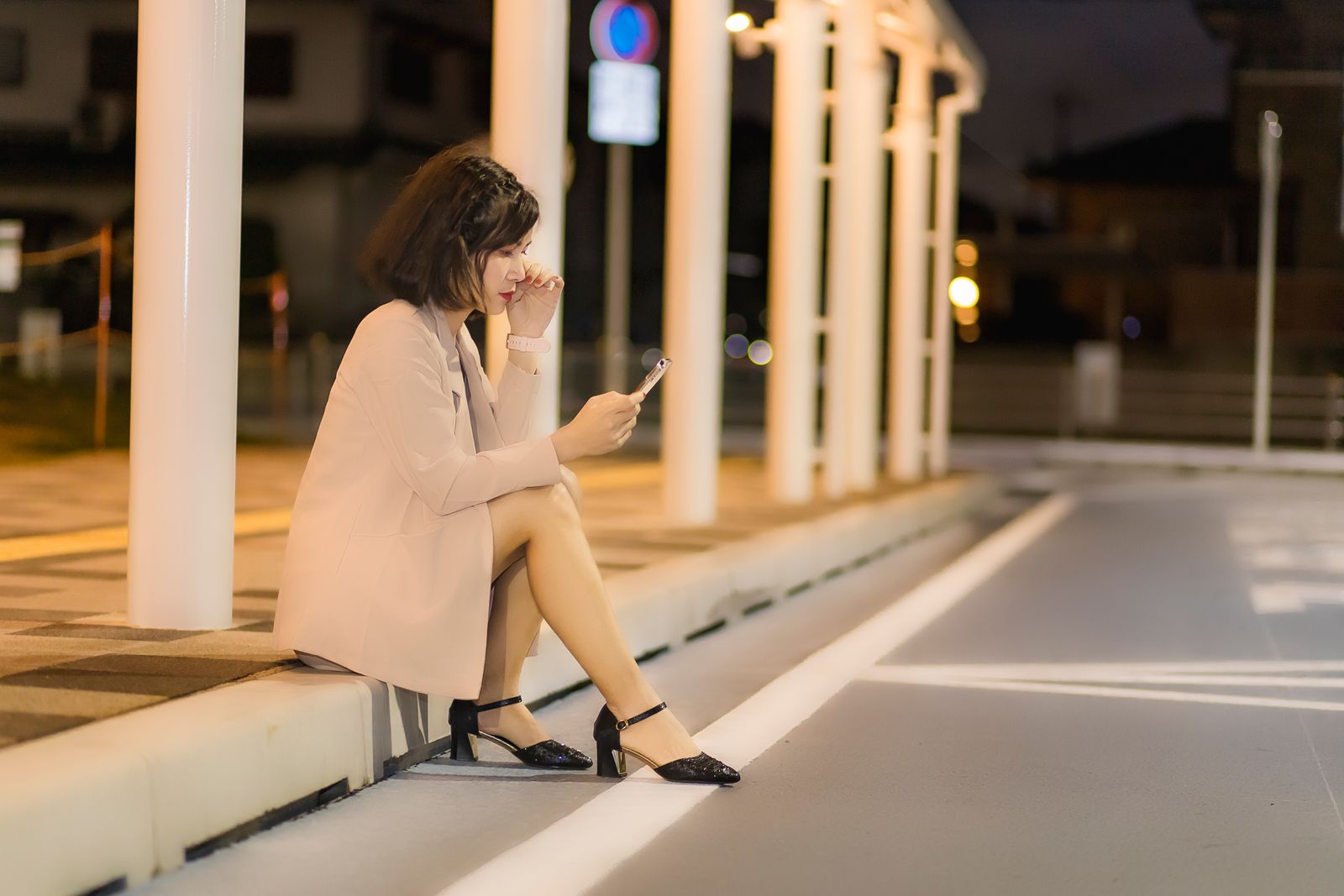 Une femme assise sur un trottoir regarde son téléphone, la nuit